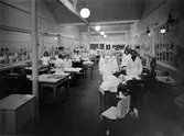 Interiörbild från Alingsås Linnefabrik AB, då Sveriges Förenade Linnefabrikers AB. Ett antal arbetare står vid sina arbetsbord.