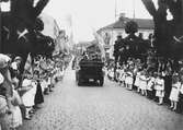 Kung Gustav V:s besök i Borås 1924. Ett ekipage med bilar färdas över den utsmyckade Västerbron på Stora Brogatan i riktning mot stadskärnan. Barn med flaggor står längs med vägen.