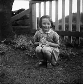 Lilla Ann-Sofi Ottosson på Filialen (butik) i Fageråkra sitter med en kattunge i knät invid ett staket, i hörnet mot Evert Petterssons hus.