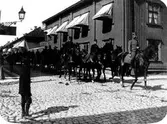 Gatuvy av en hästparad, längst fram en man i uniform och bakom sig två rader av män, som rör sig genom standen.
A2 rider genom Alingsås, här på Kungsgatan med kvarteret Pärlan 7 i bakgrunden.