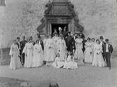 Brudpar med bröllopsgäster framför en pampig barockportal. Brudgum och några gäster bär uniformer.