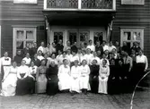 Gruppbild med kvinnor och ett fåtal män, varav en ser ut att bära prästkrage, vid entrén till en träbyggnad med balkong. Ytan är stensatt och har vattenrännor, så troligen ligger huset vid en stadsgata.
