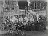 Grupp med uppklädda kvinnor och män framför en stor glasveranda med fritrappa. Kanske är det en prästgård då tre präster finns i sällskapet. De äldre damerna bär stora hattar med hakband.