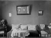 Interiör med bord vid dagbädd med stora kuddar och däröver en spegel samt två små tavlor. Textilierna har jugendmönster.