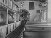Interiörbild av Torpa kyrka. Ålderdomlig inredning med predikstolen kvar på höger sida om den bevarade korbågen. Läktare över bänkarna på vänster sida med trappa framme vid korbågen. Ljuskronan har inte elektrifierats.