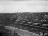 Vy från Varbergs fästning åt nordöst över den torrlagda vallgraven, societetshuset och vidare över Varberg. Den höga skorstenen hör till elkraftverket. Fotografiet är taget efter 1920 eftersom järnvägen söderut dessförinnan gick längs vägen i högerkanten.
Se även bild GB1_1099 för vyns fortsättning åt höger i bild.