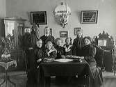 Skomakare Jacobsson med familj samlad kring ett bord i salongen.  En tramporgel står till höger om dem.