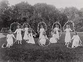 Barnlek. Tio vitklädda flickor som dansar och leker med blomsterbågar i en park.