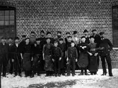 Gruppbild på 20 garveriarbetande män som står på led framför Göta läderfabrik, kv. Syrenen.