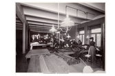 Interiör från Nordiska Kompaniets syateljé vid Stureplan, fyra trappor upp. Sömmerskor syr, skär till tyg och arbetar med olika moment vid avlånga träbord. Armaturer med glaskupor. I förgrunden ett tillskuret plagg. Redan 1902 fanns elektriska symaskiner i ateljén, elekrisk belysning installerades 1910.