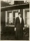 En medelålders kvinna i klänning står utanför ett mindre bostadshus. Växjö, ca 1930.