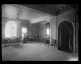 Interiör från Franska damskrädderiet på Nordiska Kompaniet, våningsplan fem. Rummet är elegant möblerat  med stilmöbler och en stor spegel med förgylld ram. Till vänster i bild ett stort fönster med välvd överdel, där solen strålar in. Ett tygsjok ligger draperat över en stol. Tjock heltäckningsmatta.