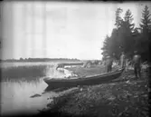 Tre män vid en uppdragen eka på en stenig strand. I bakgrunden dricker en ko vatten ur sjön.