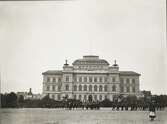 Växjö högre allmänna läroverk, ca 1905. Ett antal pojkar är ute och marscherar m.m. på skolgården, betittade av en flicka. 
Till vänster skymtar borgmästare Bergendahls hus i hörnet Trädgårdsgatan/Parkgatan och bakom det Flickskolan. 
Till höger skymtar man bokhandlare Quidings hus på Södra Järnvägsgatan.