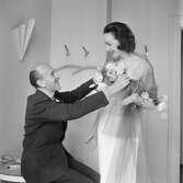 En man och en kvinna i ett provrum. Pelle Lundgren provar en vit klänning med Havannabrun tyllsjal på Hjördis Genberg Niven, före detta stjärnmannekäng på NK:s Franska damskrädderi, när hon återvänder till ateljén våren 1953.