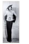 Mannekäng visar festklänning i svart och vitt med rynkat liv med tunn snodd i midjan. Matchande bolero  med pälsdetaljer. Hatt. NK:s Franska damskrädderivåren 1938.