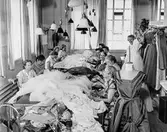 Interiör från ateljén på NK:s Franska damskrädderi. Vid arbetsbordet som är fyllt med tyger, bland annat tyll, sitter sömmerskor och arbetar med klänningar inför Amaranterbalen 1956. Proverskan Wivan Nilsson längst till höger i bild. På en ställning i rummet hänger plagg på galgar. Från taket hänger många lampor.