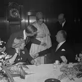 Christian Diors besök på NK, Nordiska kompaniet i Stockholm, februari 1957. Han bjuds på en visning av plagg uppsydda av tyger från NK:s textilkammare av Kurt Jacobsson och Pelle Lundgren. Mannekäng visar plagg. Vid bordet sitter Dior tillsammans med Astrid Sampe.