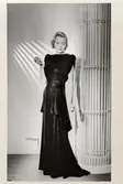 Mannekängen Margit Langlet poserar iklädd hellång, svart sammetsklänning med kort, draperad ärm och skört, sedd framifrån. Från NK:s Franska damskrädderis egen ateljé. Klänningen bakifrån se NMA.0099258.