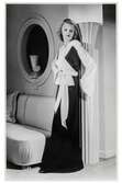 Mannekängen Margit Langlet visar festplagg i provrum med klassicistisk dekor, NK:s Franska damskrädderi. Aftonklänning, hellång, av vit och svart crêpe, med långa vida ärmar. Brett knytskärp i rosett markerar midjan.