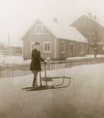 En pojke poserar på sparkstötting framför ett bostadshus. Okänd ort, ca 1910.