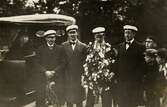 Tre ungherrar i rock och studentmössa uppvaktar en fjärde herre på hans studentdag. Till vänster väntar en kvinna i en bil och till höger beskådas det hela av fyra nyfikna pojkar. Ca 1920.