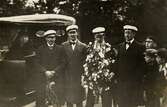 Tre ungherrar i rock och studentmössa uppvaktar en fjärde herre på hans studentdag. Till vänster väntar en kvinna i en bil och till höger beskådas det hela av fyra nyfikna pojkar. Ca 1920.