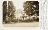 Ett större bostadshus med veranda, i en trädgård. Ca 1908. 
Man skymtar en kvinna och en man på verandan.