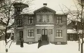 Ett bostadshus i snö, med två kvinnor stående i trädgården framför. Ca 1914.