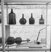 Utställning Tiotusen år. Glasmonter i utställningsdelen Bondesamhället 1800- talet. På översta hyllplanet Brännvinsflaskor i olika storlekar. På nedre hyllplanet ligger en fiol med nothäfte, pipa med en tobakspung och ett fickur.