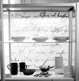 Utställning Tiotusen år. Glasmonter i utställningsdelen Bondesamhället från 1800-talet. På översta hyllplanet Porslintallrikar, på nedre hyllplanet stop av lergods, bestick och skålar.