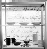 Utställning Tiotusen år. Glasmonter i utställningsdelen Bondesamhället från 1800-talet. På översta hyllplanet Porslintallrikar, på nedre hyllplanet stop av lergods, bestick och skålar.