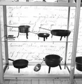 Utställning Tiotusen år. Glasmonter i utställningsdelen Bondesamhället från 1800-talet. I montern finns det på översta planet kittel och stekpannor, åderlåtningsjärn, rakniv och tandborste. På det nedre planet finns det Stekpanna och gryta med slev.