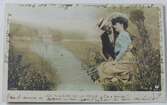 Vykort med romantiskt motiv, en man och kvinna sitter vid en bäck, fiskar och omfamnar varandra. Den franska handskrivna texten som täcker baksidan fortsätter även på framsidans kanter.