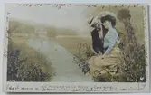 Vykort med romantiskt motiv, en man och kvinna sitter vid en bäck, fiskar och omfamnar varandra. Den franska handskrivna texten som täcker baksidan fortsätter även på framsidans kanter.
