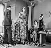 Mannekäng poserar i hellång blommig klänning i provrummet vid NK:s Franska damskrädderi. I soffan och på ett bord sitter damer, presumtiva kunder. På golvet ligger tyg. Pelle Lundgren står vid sidan.