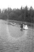 En man som står i vattnet och placerar ut en modellbåt. I bakgrunden en roddbåt.
