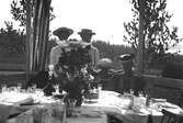 Ett dukat bord prytt med blommor. Bakom bordet står det två kvinnor och en pojke. Till höger en sittande kvinna.