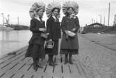Tre flickor som står på kajen invid Skeppsbron. Flickorna har små korgar som de håller i sina händer.; 
