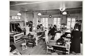 Beklädnadsateljéerna på NK (Nordiska Kompaniet). I lokalen arbetar både män och kvinnor med sömnadsarbeten vid långa bord. I förgrunden står två symaskiner.