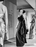 Mannekänger visar festplagg framför spegel i provrummet. Den ena kvinnan bär hellång mönstrad klänning, figurnära med axelband, drapering vid stussen. Den andra kvinnan bär en sammetsklänning av liknande snitt. Runt halsen ett fyrradigt pärlcollier. NK:s Franska damskrädderi.