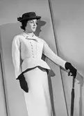 Mannekäng visar plagg. Dräktjacka, figursvängd, höftlång och dubbelknäppt med pennkjol. Hatt, handskar och paraply i ena handen. NK:s Franska damskrädderi.
