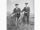 Ateljéfoto. Två uniformerade ynglingar vid var sin cykel mot havsfond. Troligen cykelbud.