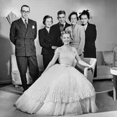 Friherrinnan Susanne Palmstierna poserar i den Diorklänning hon vunnit vid utlottning på NK:s Franska damskrädderi. Den ärmlösa klänningen har plisserat liv och kjol, midjan markerad med skärp. Underkjol av plisserad tyll. Bakom henne står juryn med bland andra Kurt Jacobsson.