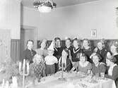 Julfest för 8:e klass i Flickskolan i ett julpyntat rum med väggbröstning. Ett litet barn deltar också och alla är samlade i små partyhattar vid bordsänden. (Se även bild GB2_3679)