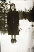 Anna Snygg står på en snöig skogsväg med hatten långt bak på huvudet, skinnhandskar och pälsbrämade stövletter.