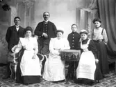 Sju personer på en ateljébild; fyra kvinnor, två män och en pojke. Troligen är de tjänstefolk hos någon familj.