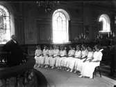 Varbergs kyrka. Komfirmandgrupp bestående av vitklädda flickor. Gruppen sitter på stolar i kyrkans kor. Innanför altarringen står prästen. I kandelabrarna sitter elektrifierade ljus.