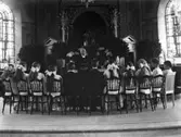 Konfirmation i Varbergs kyrka år 1915. Konfirmanderna sitter på stolar kring altarringen. De har ryggen vänd mot kameran. Lägg märke till de elektrifierade 
