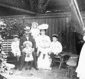 Familjebild av makar med fem barn på en trädgårdssoffa. Det kan vara byggmästare Johannes Nilsson med familj. Till höger i bild syns troligen en assistent till fotografen som håller upp ett skuggande skynke.